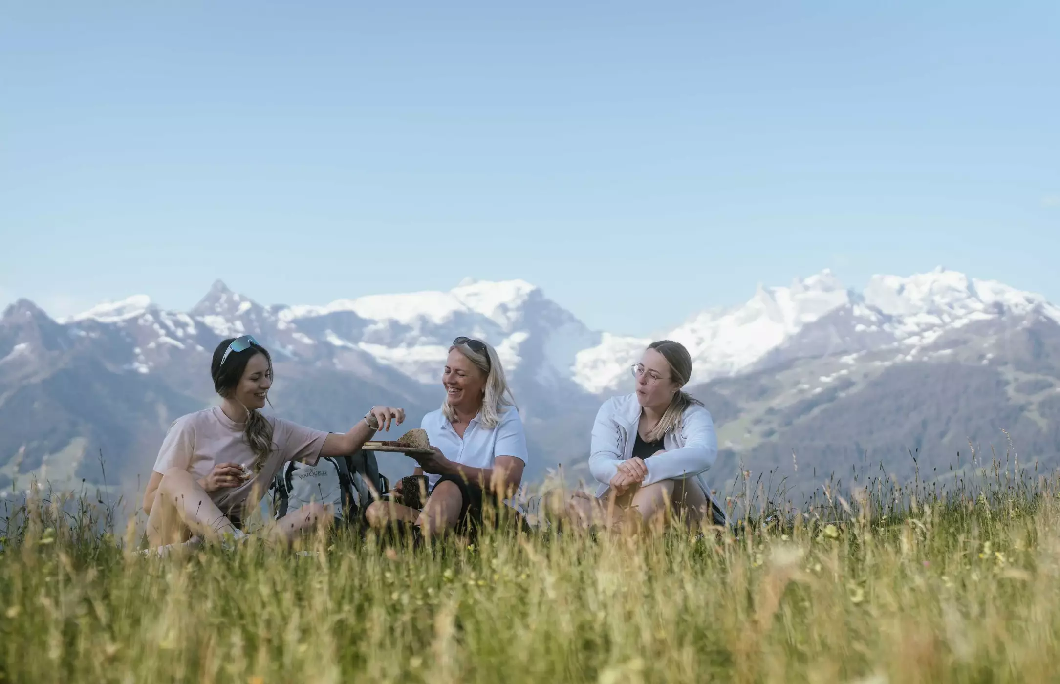 Drei Frauen sitzen auf einer Wiese in den Alpen, genießen ein Picknick und lachen, im Hintergrund schneebedeckte Berge.